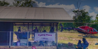 Image of remote AEC Polling Place. Staff are setup in a shed, members of the public area seated in the foreground with a couple of dogs, and a helicopter is in the background. 